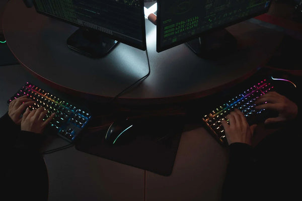 Two cybersecurity professionals typing on RGB backlit keyboards while monitoring computer screens with code in a dark workspace.