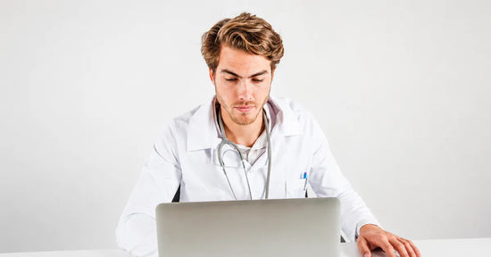 Young male doctor in white coat working on a laptop in a minimalist studio.