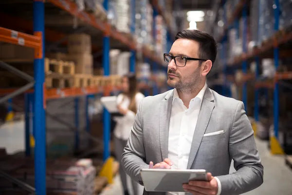 A business professional wearing glasses and a suit holds a tablet while inspecting operations inside a warehouse with stocked shelves in the background.